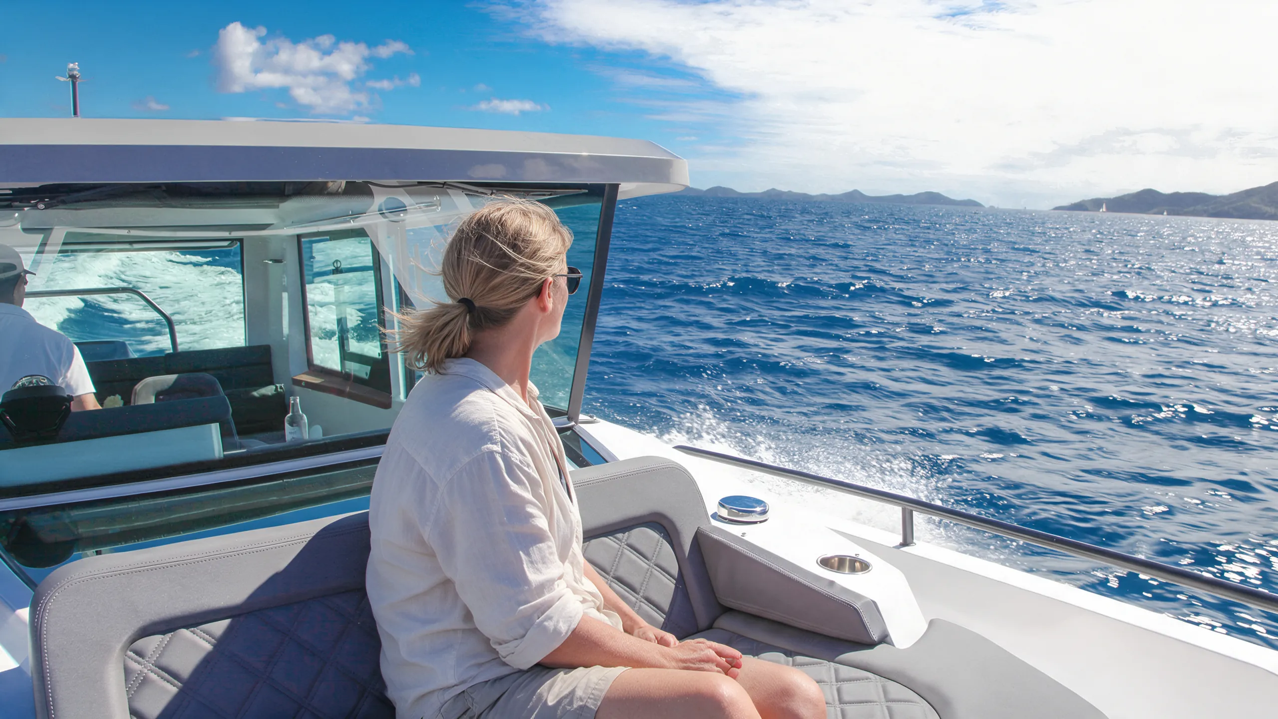Passenger with hair tied back looking out at the sea from the cockpit of a water taxi underway in the British Virgin Islands, with the captain at the helm and green island hills on the horizon