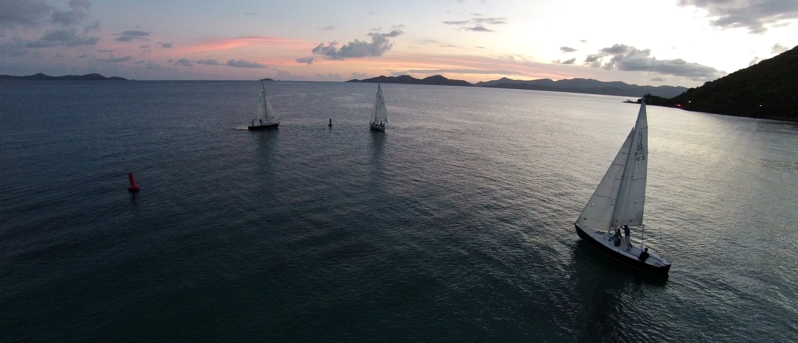 Sailboats gliding across calm water at dusk under a pink and violet sky, with island silhouettes on the horizon in the Virgin Islands