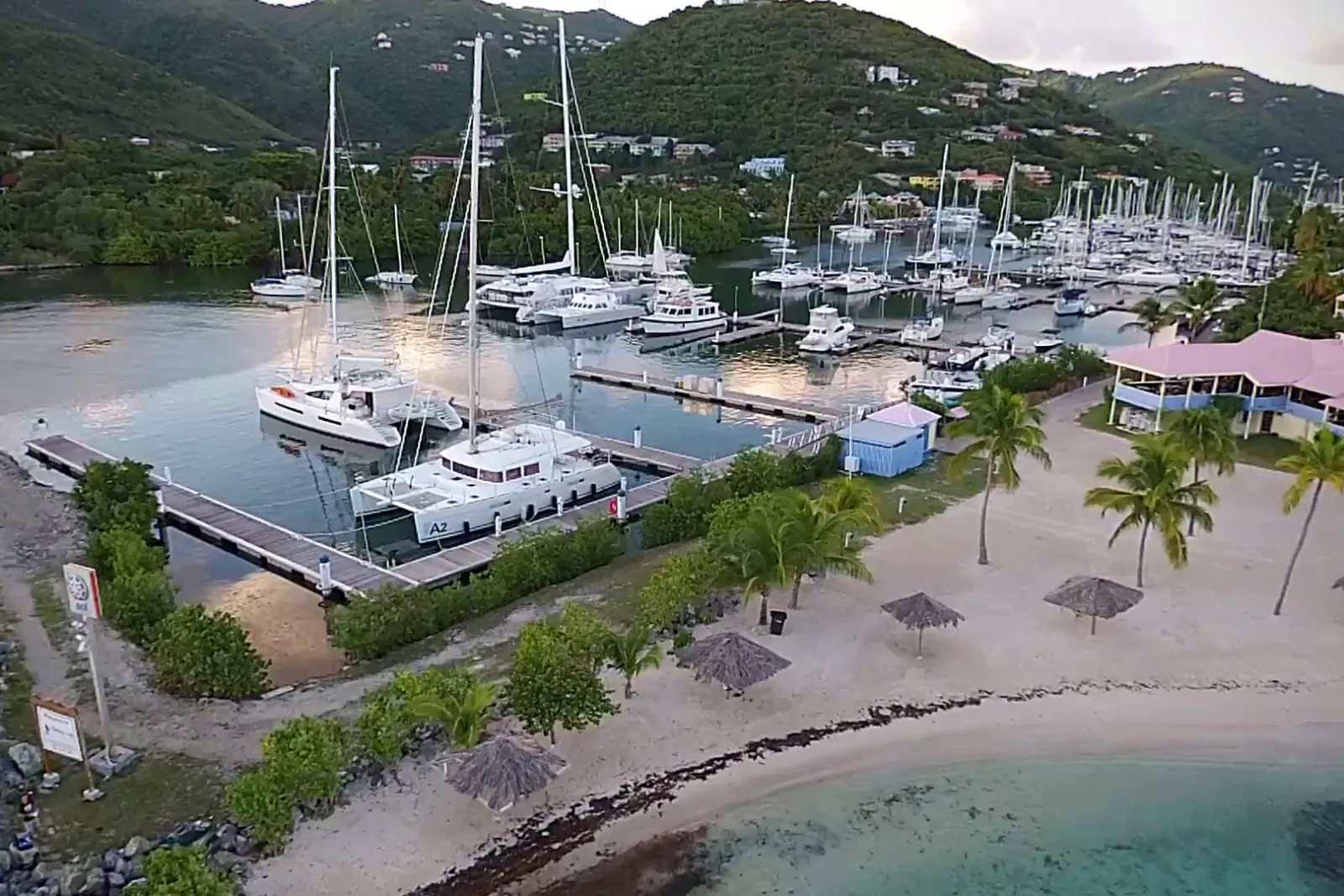 Aerial view of catamarans and sailing yachts berthed at Nanny Cay marina, Tortola, in soft evening light, with a palm-lined beach in front